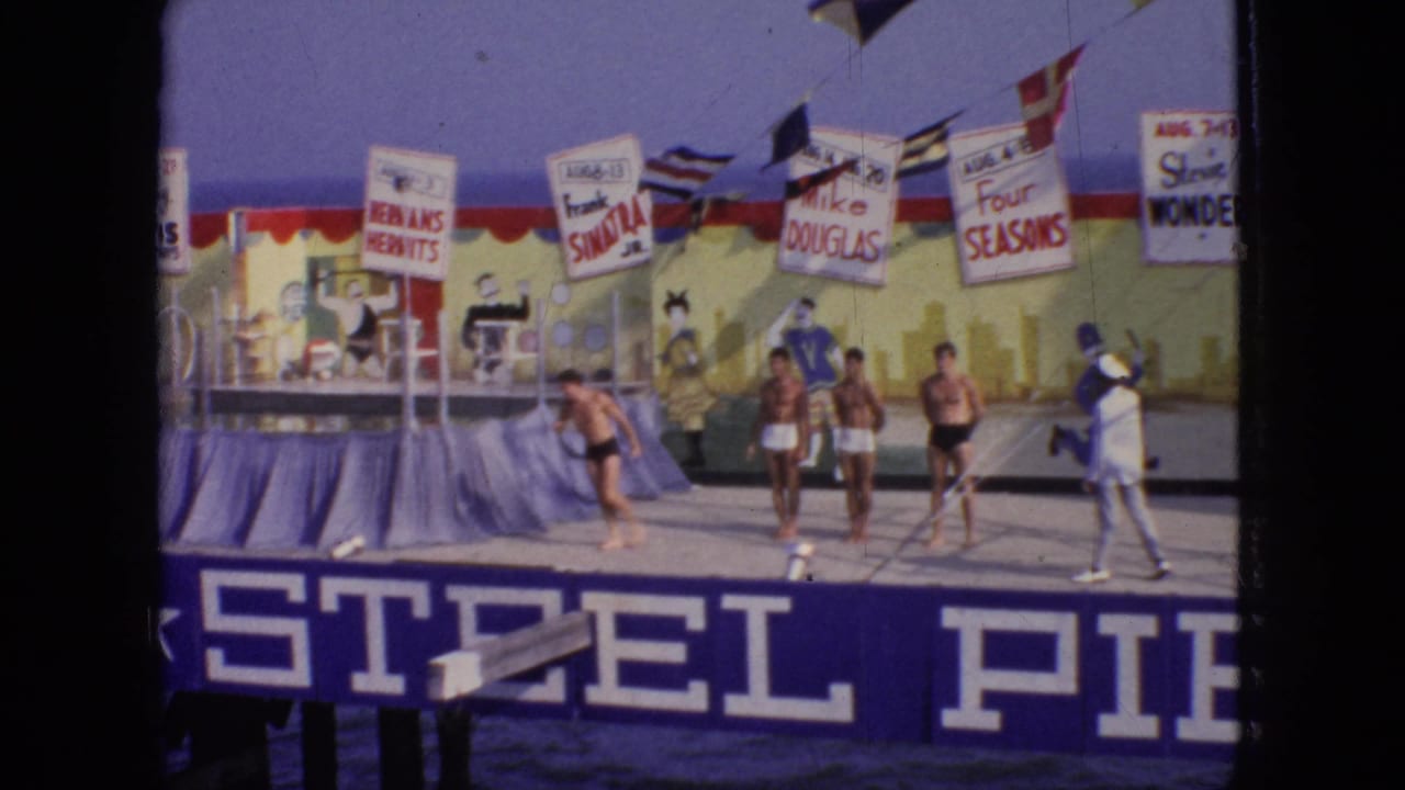 1966 New Jersey frame showing young men in swim trunks on the Steel Pier stage, with flags, performer-name placards, and the painted backdrop visible behind them.