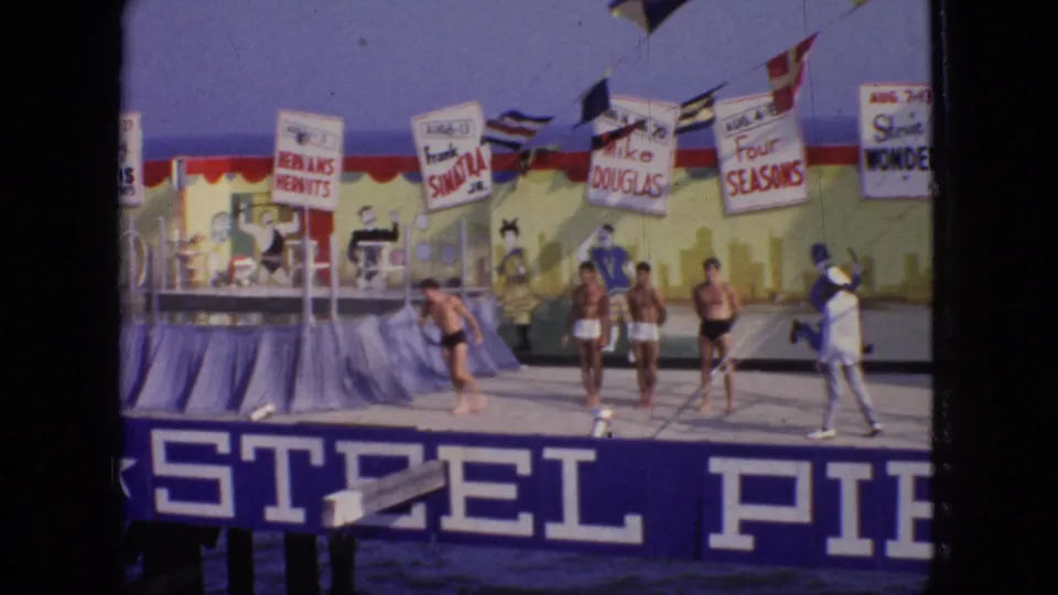 1966 New Jersey frame showing young men in swim trunks on the Steel Pier stage, with flags, performer-name placards, and the painted backdrop visible behind them.