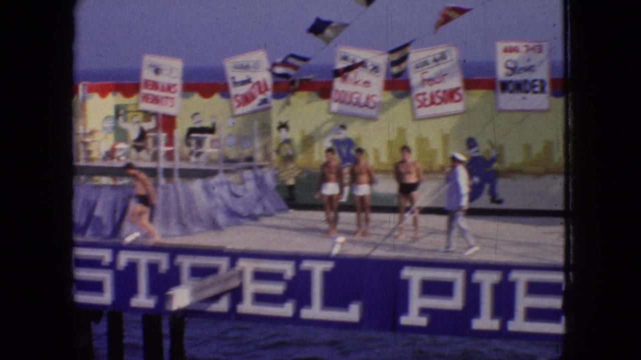 A 1966 New Jersey home-movie frame shows a Steel Pier performance stage with several people near the front, show banners overhead, and large pier lettering below.