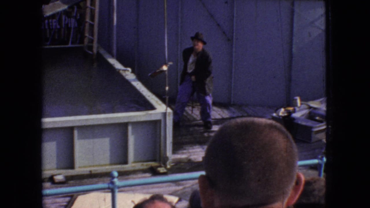 Male performer in a hat stands beside a raised stage area as audience heads fill the foreground.