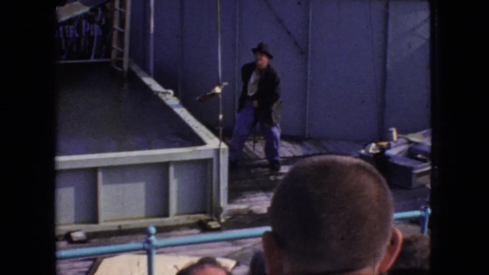 A male performer in dark jacket, hat, and blue trousers stands near the edge of a Steel Pier stage while audience members are visible in the foreground.
