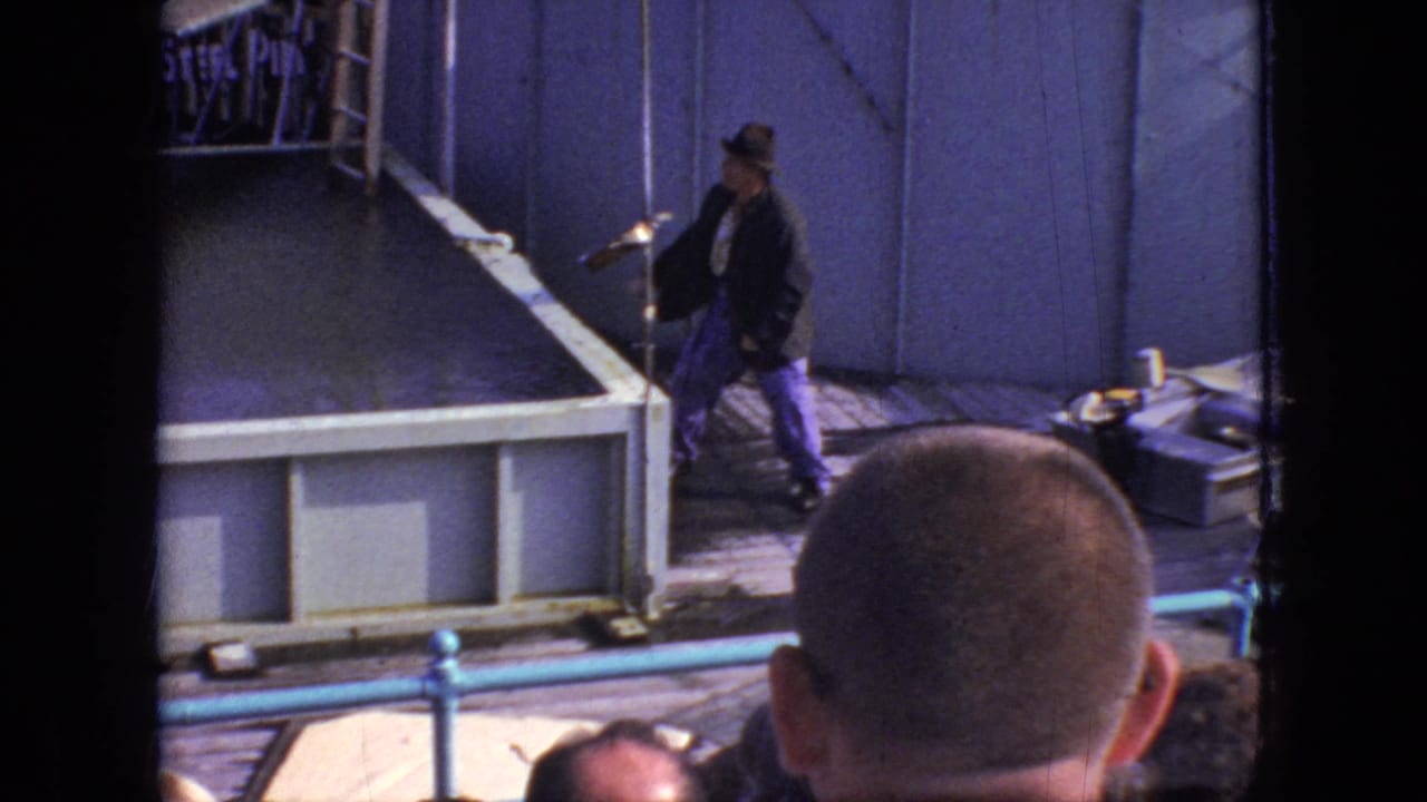 The handheld frame catches the performer beside the blue stage wall and padded platform, partly blocked by the head of a nearby spectator.