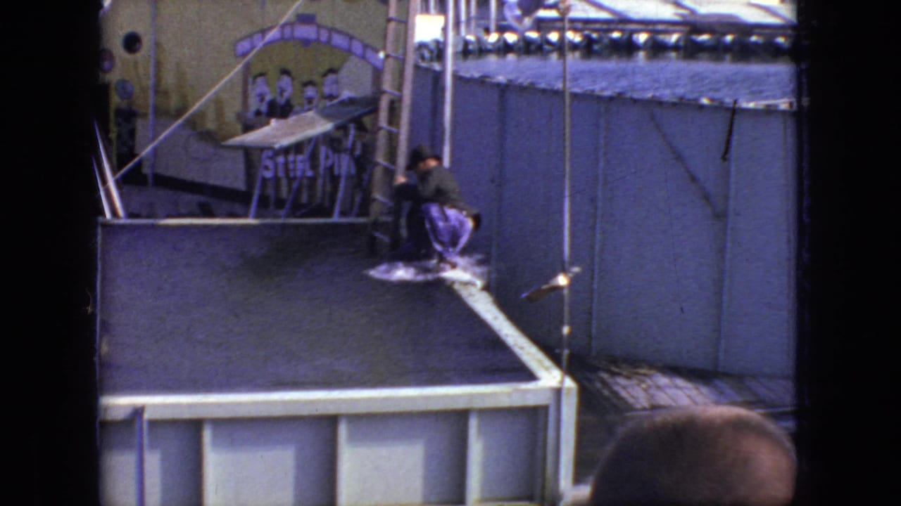 At the end of the 1966 Steel Pier stunt moment, the performer appears low on or near a padded landing surface beside the stage, with audience heads at the bottom edge.