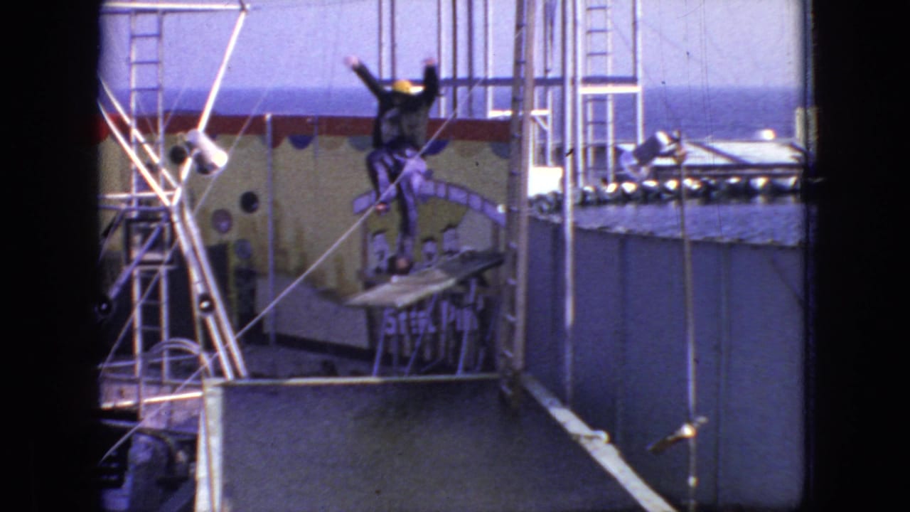 In a 1966 New Jersey 8mm home-movie frame, a person in a purple outfit and yellow cap stands with arms raised on a small platform above a raised pool while metal rails and a painted backdrop sit behind them.
