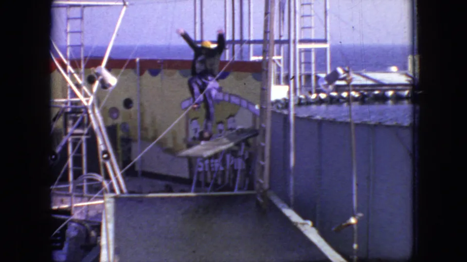 In a 1966 New Jersey 8mm home-movie frame, a person in a purple outfit and yellow cap stands with arms raised on a small platform above a raised pool while metal rails and a painted backdrop sit behind them.
