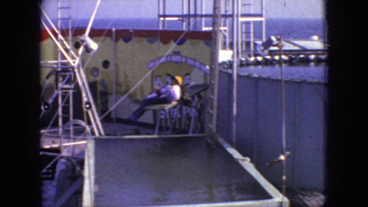 The person in the purple outfit appears seated or reclining on the diving platform beside the raised pool, with metal framework, decorative backdrop panels, and onlookers visible in the 1966 New Jersey home-movie setting.