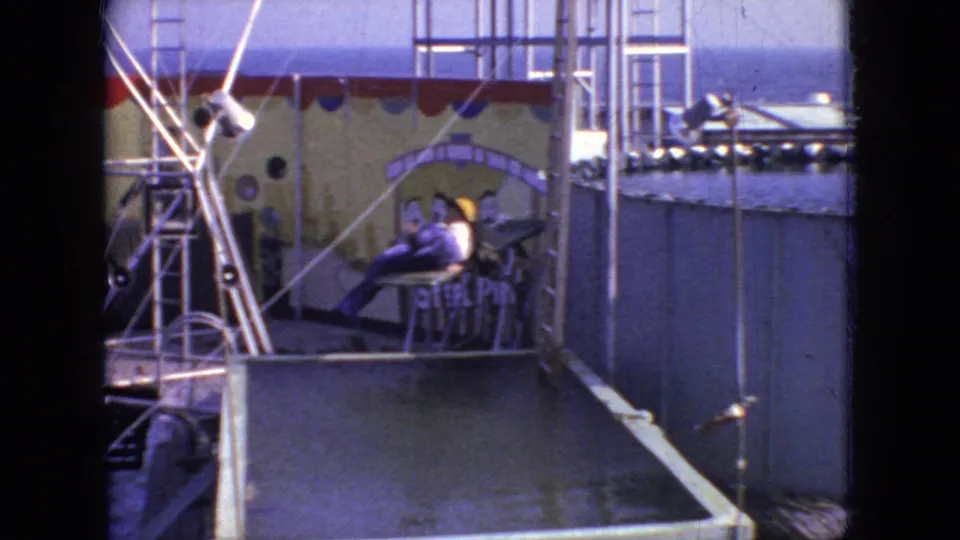 The person in the purple outfit appears seated or reclining on the diving platform beside the raised pool, with metal framework, decorative backdrop panels, and onlookers visible in the 1966 New Jersey home-movie setting.