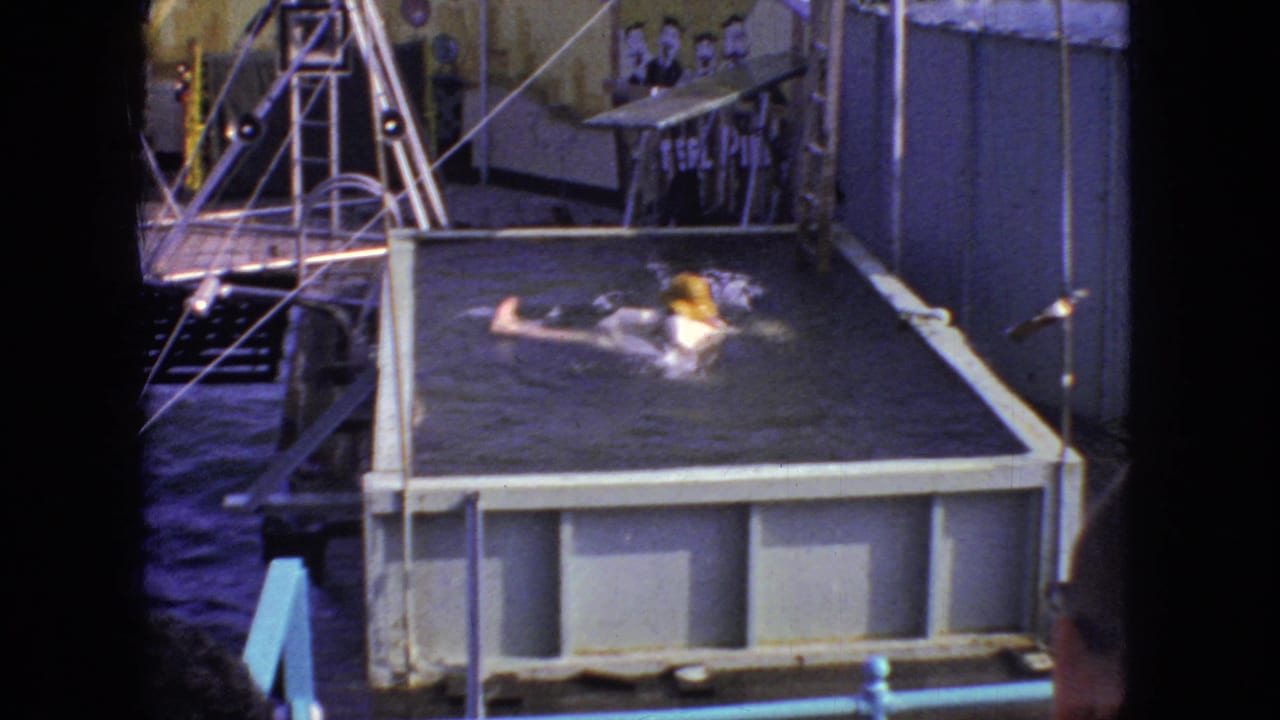 Water splashes around the person in the raised pool after the backward fall, while the small platform, metal railings, painted backdrop, and seated figures remain visible in the 1966 New Jersey 8mm frame.