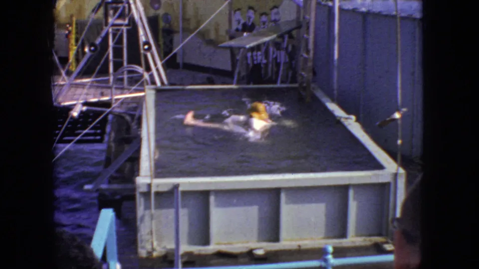 Water splashes around the person in the raised pool after the backward fall, while the small platform, metal railings, painted backdrop, and seated figures remain visible in the 1966 New Jersey 8mm frame.