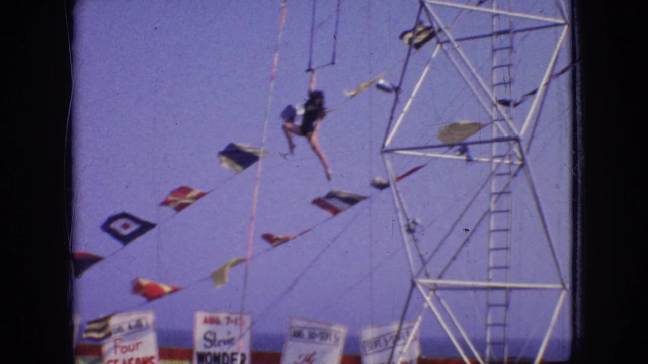 A female trapeze performer appears suspended on ropes beside a tall metal aerial rig at an outdoor New Jersey fair or circus setting in 1966.