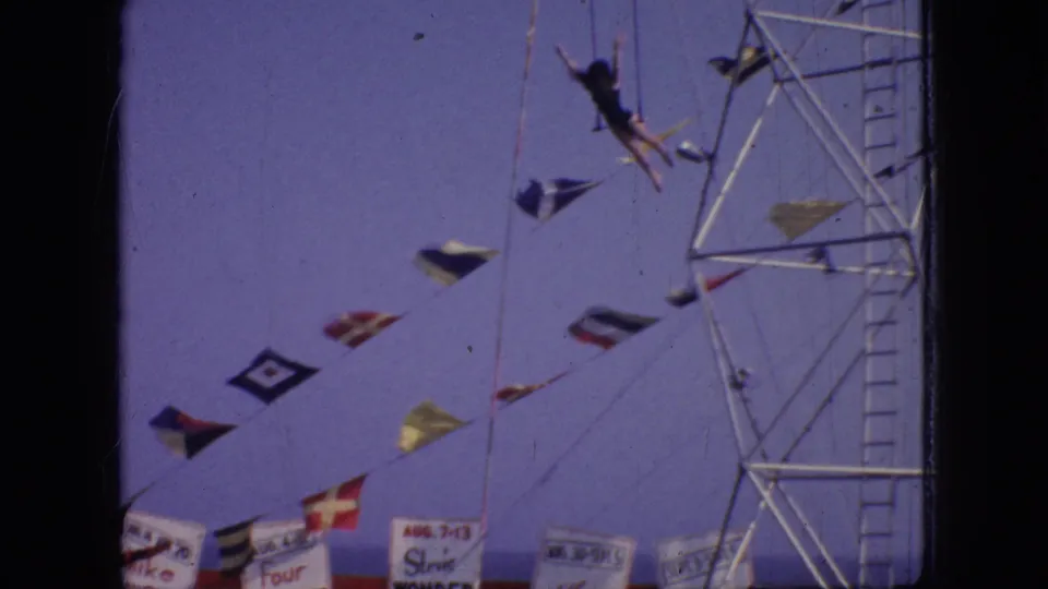 The trapeze artist swings outward from the suspended ropes, framed by strings of flags, partial banners, and the metal aerial rig.