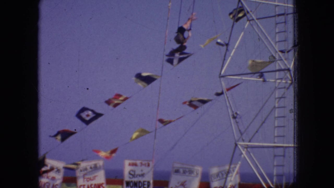 The aerial performer appears upside down during a flip or turn beside the metal rig, with flags and partial event signage below.