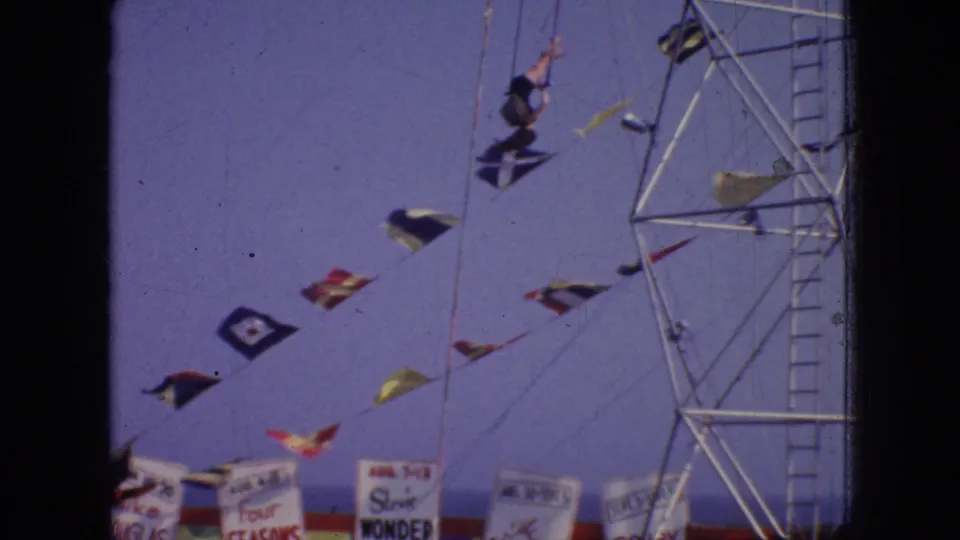 The aerial performer appears upside down during a flip or turn beside the metal rig, with flags and partial event signage below.