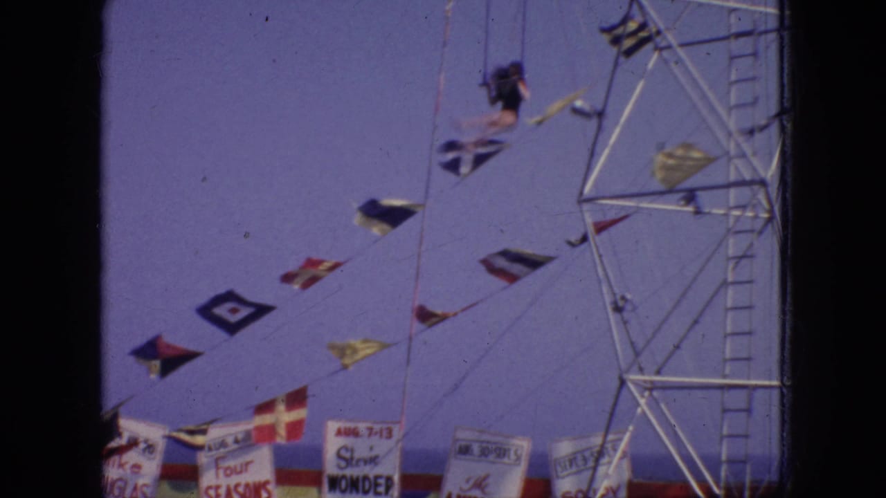 In this 1966 New Jersey home-movie frame, a dark-clad aerialist is suspended near a metal support tower above nautical flags and partially visible carnival banners.