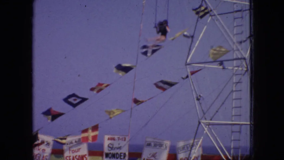 In this 1966 New Jersey home-movie frame, a dark-clad aerialist is suspended near a metal support tower above nautical flags and partially visible carnival banners.