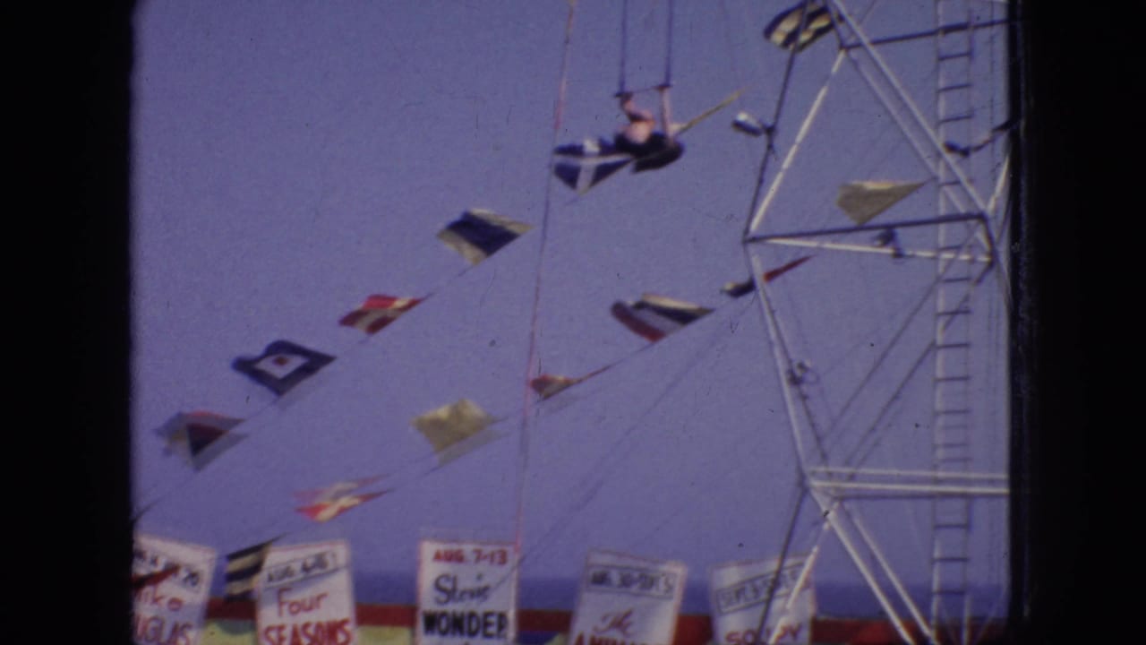 The aerialist appears stretched out near trapeze rigging, with strings of nautical flags and partially readable carnival banners below against the faded sky.