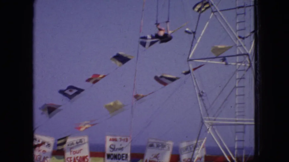 The aerialist appears stretched out near trapeze rigging, with strings of nautical flags and partially readable carnival banners below against the faded sky.