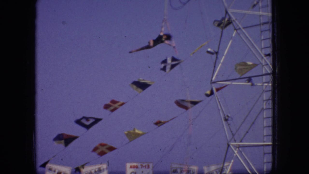 A dark-leotard aerialist hangs from the suspended apparatus beside a tall metal tower, with colorful flags and carnival signage below in the 1966 New Jersey footage.
