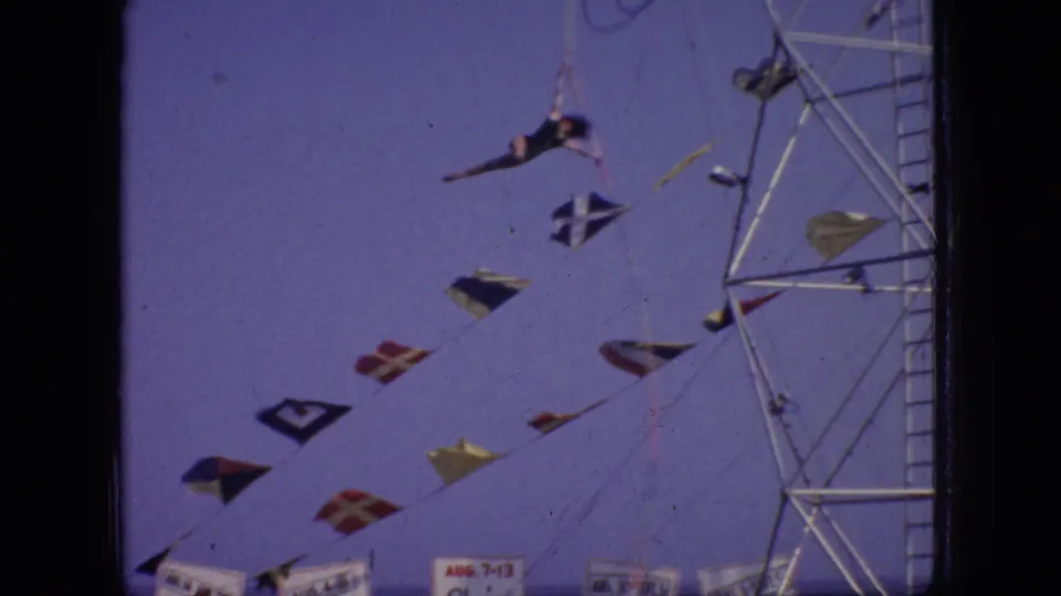 A dark-leotard aerialist hangs from the suspended apparatus beside a tall metal tower, with colorful flags and carnival signage below in the 1966 New Jersey footage.