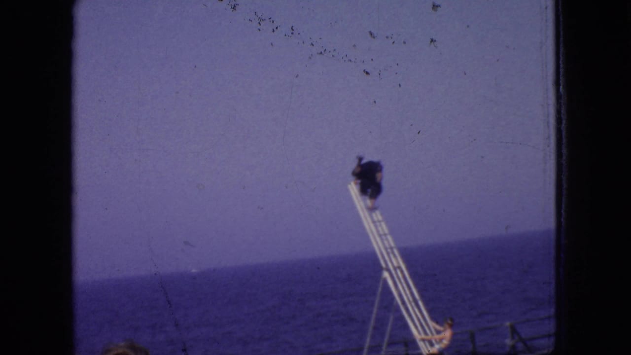 A performer appears high on a ladder or rigging structure near the seaside, with the ocean horizon and open sky visible in the background.