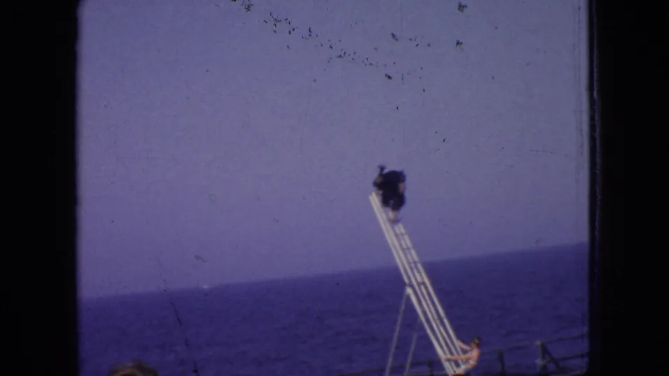 A performer appears high on a ladder or rigging structure near the seaside, with the ocean horizon and open sky visible in the background.