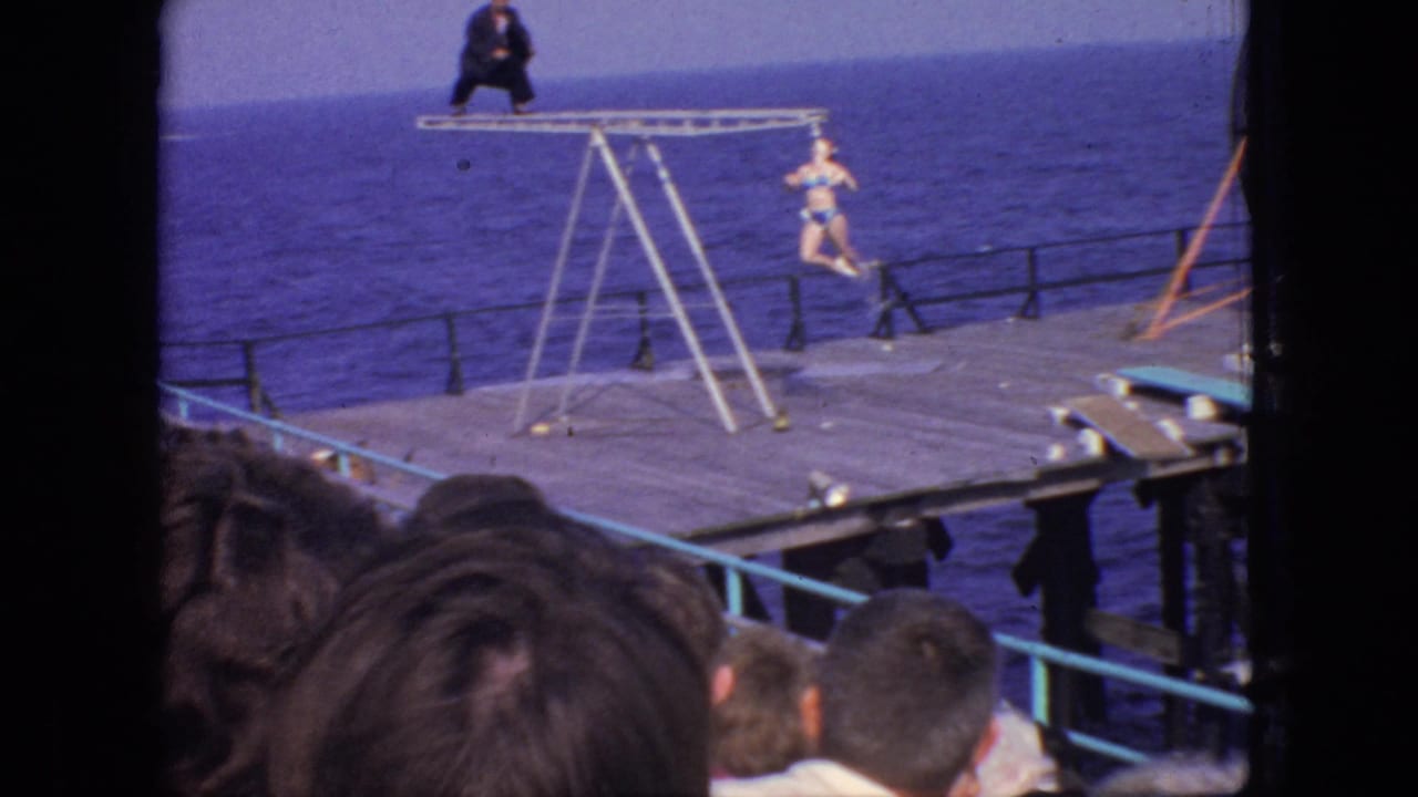 1966 New Jersey 8mm frame showing spectators in the foreground watching an acrobatic diving setup on a wooden pier with the ocean behind it.