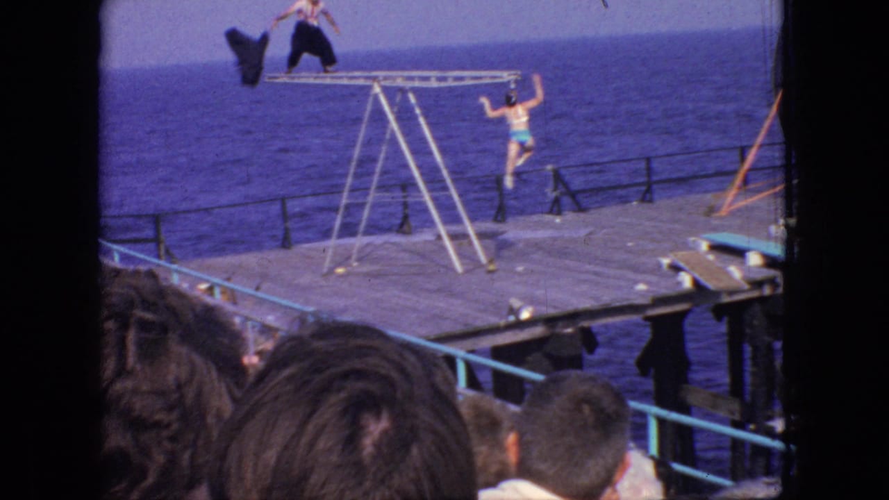 A 1966 New Jersey seaside performance frame shows a man on top of a metal ladder structure and a woman in a turquoise swimsuit on the pier below.