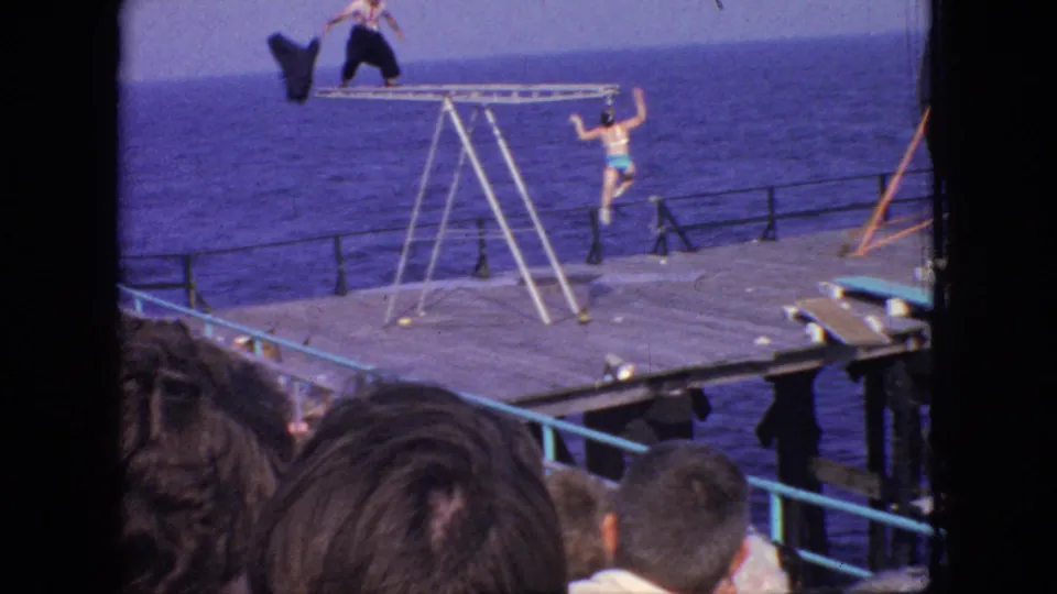 A 1966 New Jersey seaside performance frame shows a man on top of a metal ladder structure and a woman in a turquoise swimsuit on the pier below.