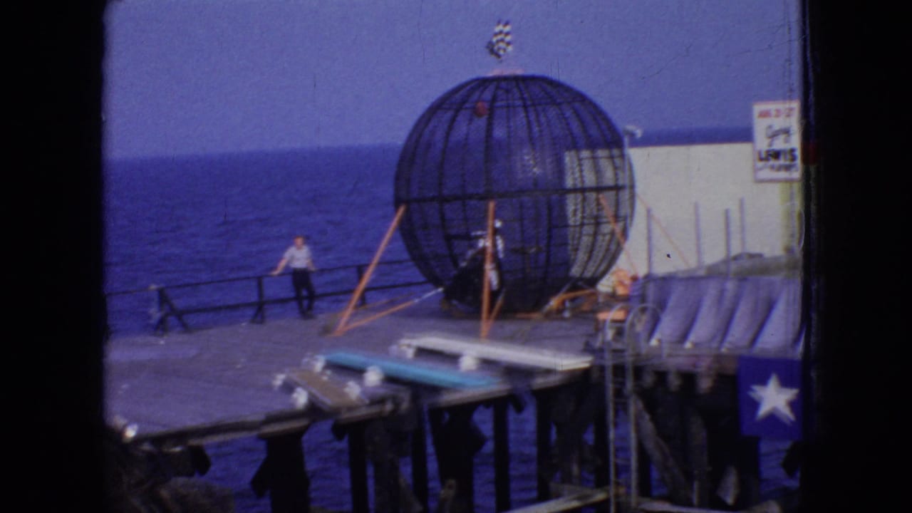 1966 New Jersey 8mm frame showing a spherical stunt cage on an oceanfront pier, with a man near the railing and signage partly visible at the edge of the frame.