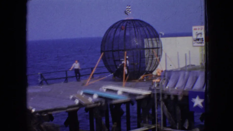 1966 New Jersey 8mm frame showing a spherical stunt cage on an oceanfront pier, with a man near the railing and signage partly visible at the edge of the frame.