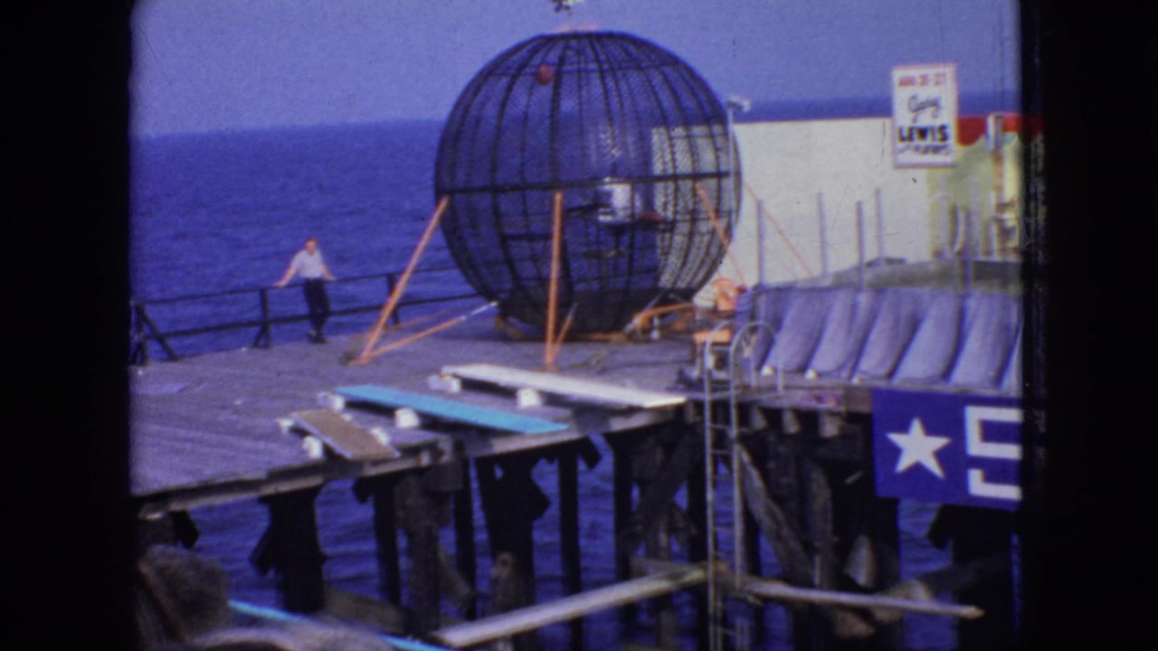 1966 New Jersey 8mm frame showing the stunt cage on the pier with a small figure visible in or near the cage, ocean beyond, and stage platforms and curtains at right.
