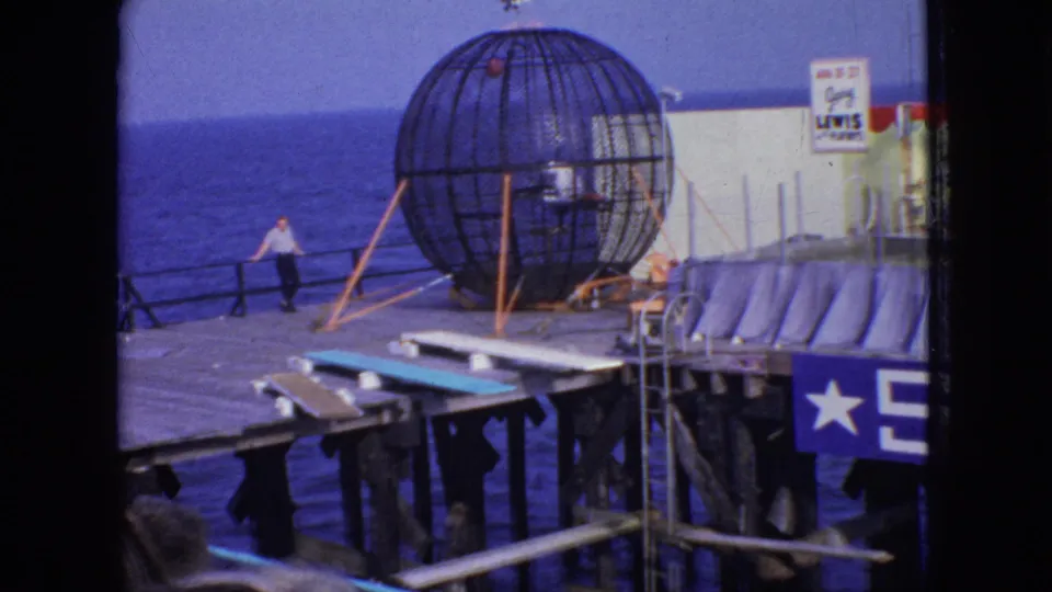 1966 New Jersey 8mm frame showing the stunt cage on the pier with a small figure visible in or near the cage, ocean beyond, and stage platforms and curtains at right.