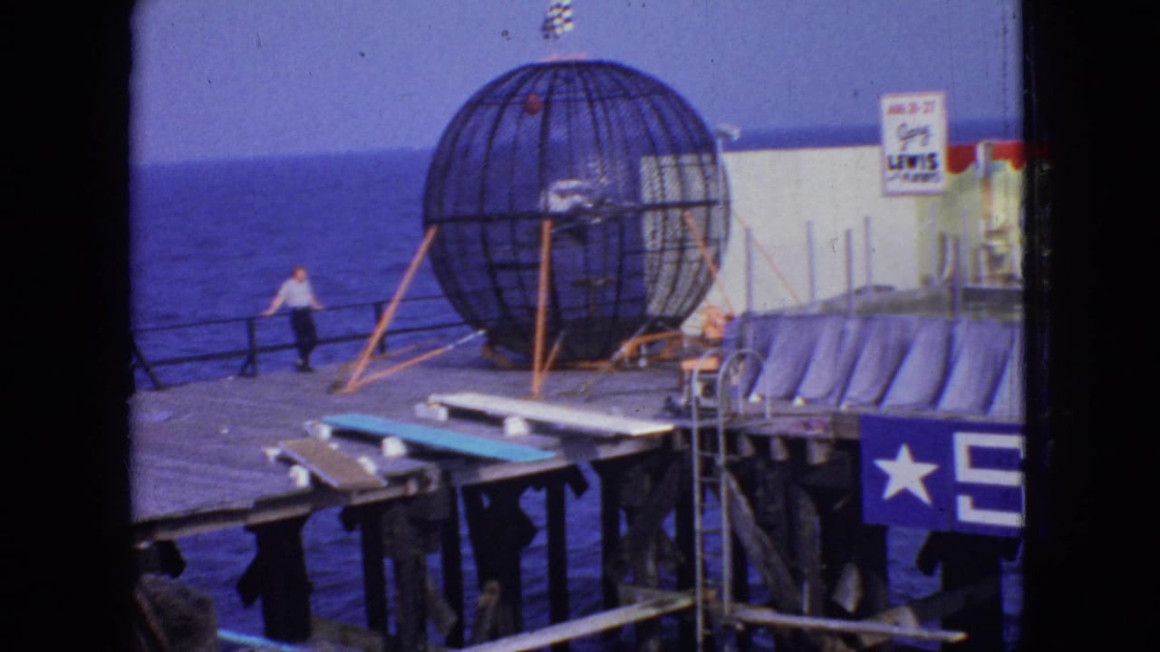 1966 New Jersey home-movie frame showing a man by the railing on a wooden pier, with a large spherical metal cage attraction and the ocean behind it.