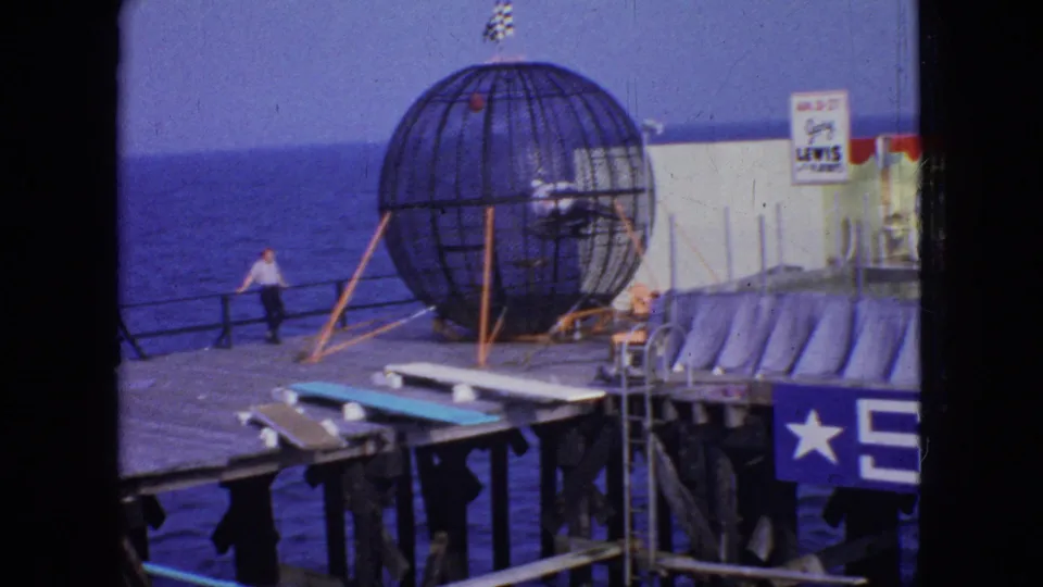1966 New Jersey pier scene with a spherical metal cage attraction, a person inside the cage, a man by the railing, and signage on the structure behind it.
