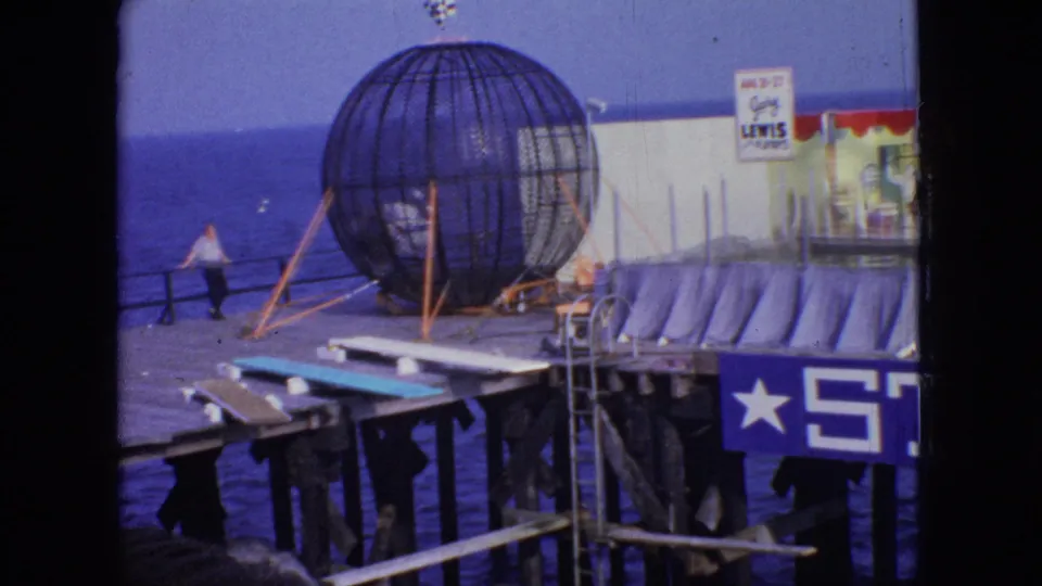 Home-movie view of a New Jersey ocean pier in 1966, showing the spherical cage attraction, a visitor near the railing, bracing poles, and partial painted signage.