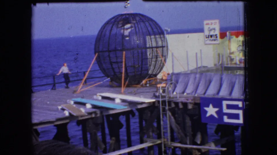 1966 New Jersey shore frame showing a large spherical metal cage on a wooden pier, a man near the railing, ocean water below, and partial amusement-pier signage.
