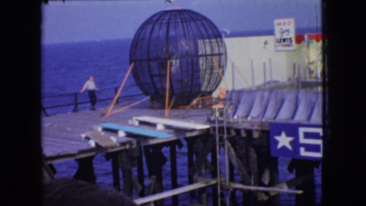 New Jersey 1966 home-movie frame showing a wooden seaside pier, ocean water, a large steel globe cage, and a man in a light shirt near the railing.