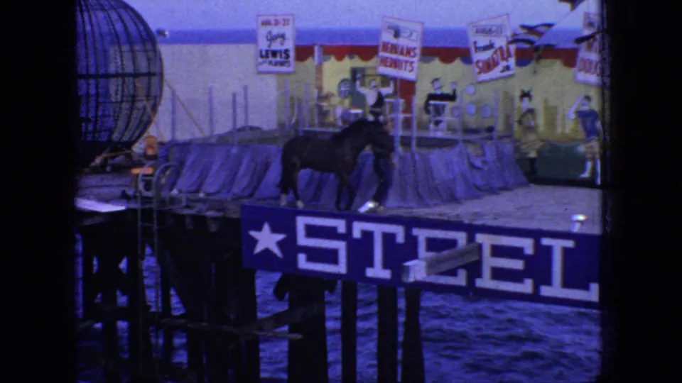 New Jersey 1966 pier circus frame showing a dark horse moving on the deck near handlers, show signs, and a large partial pier sign above the water.