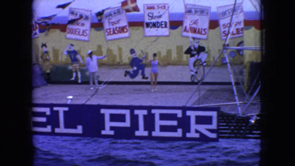 New Jersey 1966 home-movie frame showing performers on a pier stage beneath signs for circus-style acts, with a large pier sign and water below.