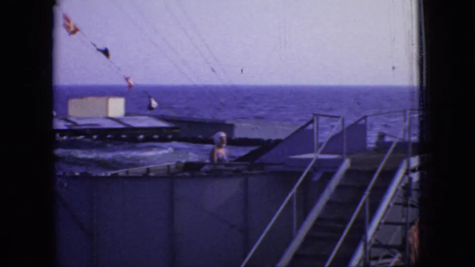 The camera view drops to the oceanfront landing area, showing bunting flags, railings, stairs, and a visible person near the pool or tank edge in the 1966 New Jersey setting.