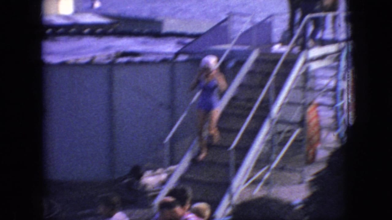 A woman in a swimsuit moves down the metal stairs from the deck area, with bystanders below, railings in the foreground, and the waterfront setting still visible.