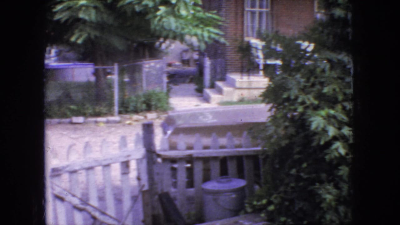1966 New Jersey residential exterior with steps, railing, fence, dense greenery, and a trash can visible from behind a dark window frame.