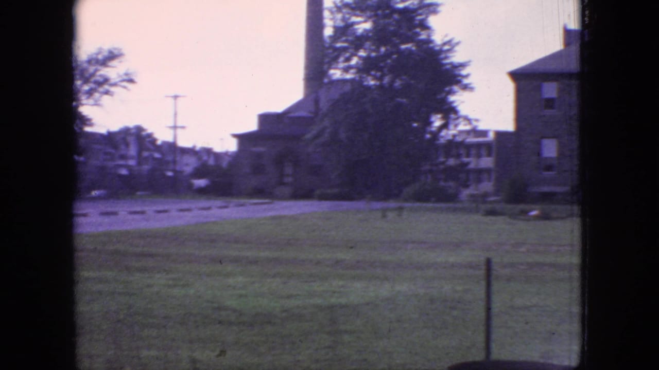 1966 New Jersey 8mm frame showing grassy grounds, a road or drive, brick buildings, utility poles, trees, and a dark window edge.