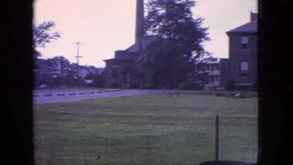 1966 New Jersey 8mm frame showing grassy grounds, a road or drive, brick buildings, utility poles, trees, and a dark window edge.