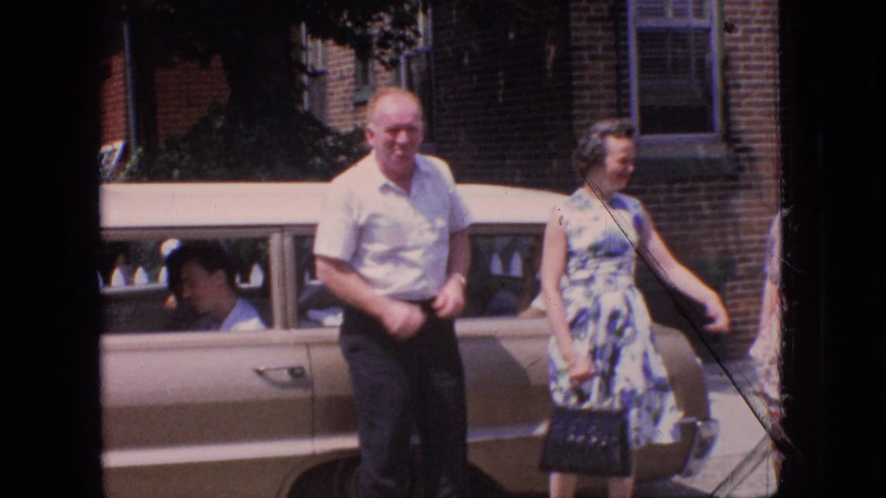 In this 1966 New Jersey home-movie frame, an older man in a white short-sleeved shirt and a woman in a blue-and-white floral dress stand beside a tan sedan with a seated person visible inside.
