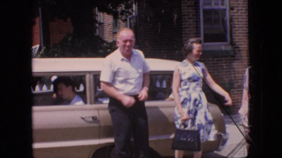 In this 1966 New Jersey home-movie frame, an older man in a white short-sleeved shirt and a woman in a blue-and-white floral dress stand beside a tan sedan with a seated person visible inside.