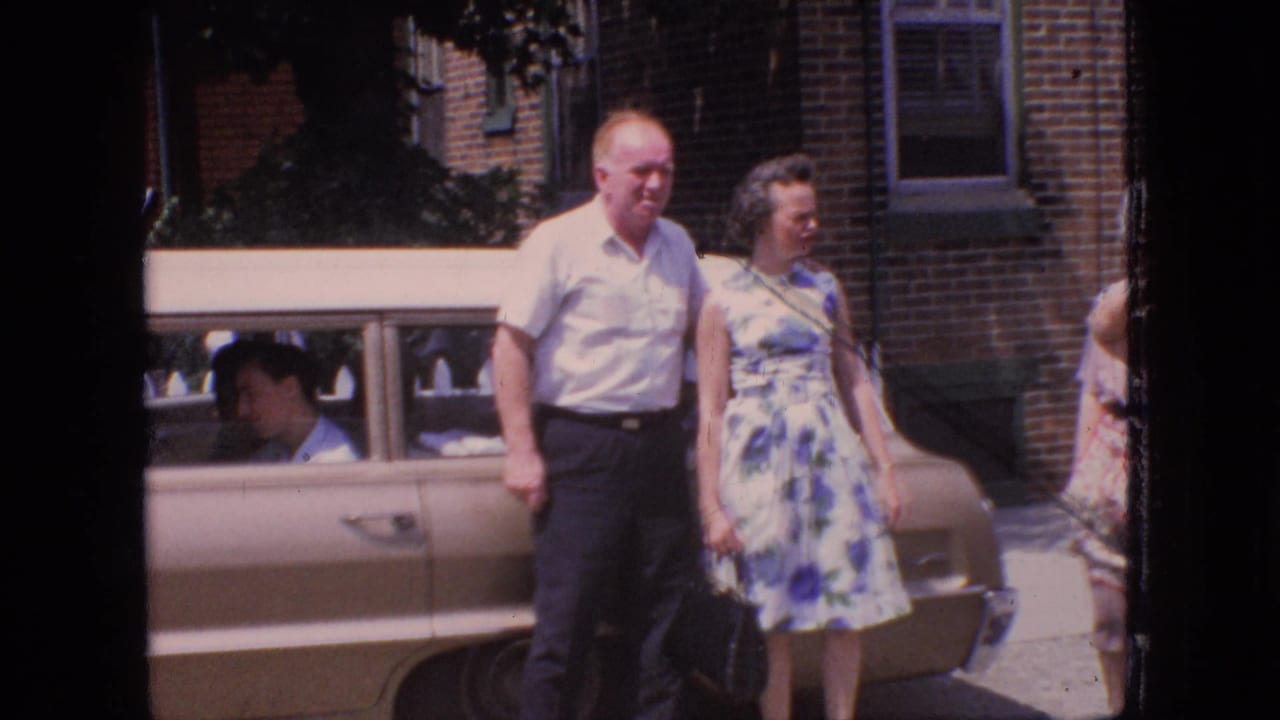 Older couple standing near a tan sedan with brick buildings behind them.