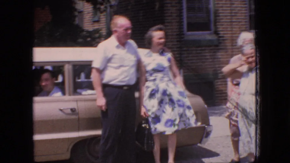 The 1966 home-movie view shows the couple standing next to the sedan while another older woman in a light dress appears at the right edge, suggesting a casual group gathering.