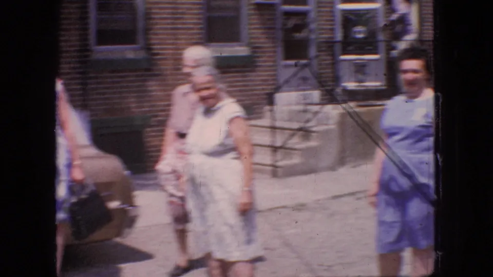 Several women move through the frame near the rear of the tan sedan, with brick residential steps and sidewalk visible in the background in the supplied New Jersey 1966 setting.
