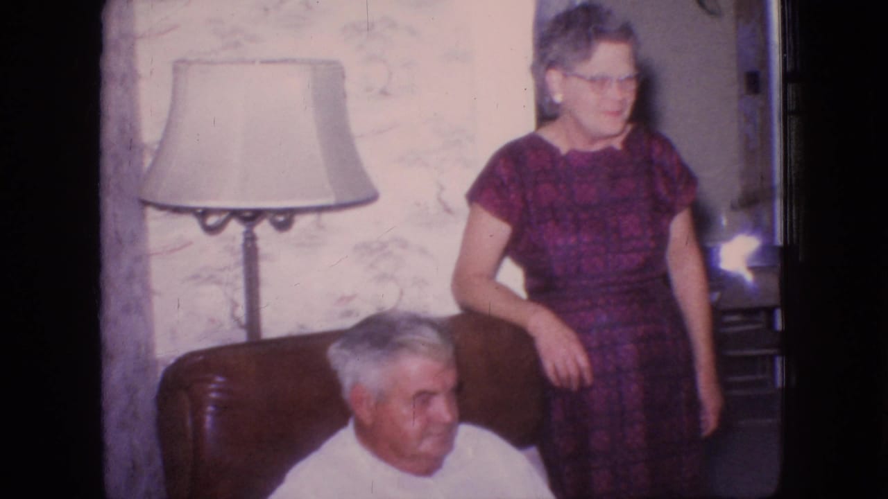 In a 1966 New Jersey home-movie frame, an older man in a white shirt sits in a brown armchair while an older woman in a purple patterned dress and glasses stands nearby beside a floor lamp and floral wallpaper.